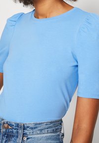 Světle modrý vršek s krátkými rukávy a kulatým výstřihem, s nabíranými detaily na ramenech, vyrobený z měkké látky, spárován s džíny s vysokým pasem.