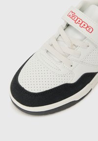 Hvit sneaker med svarte semskede detaljer, perforert design på tåen og borrelås. Har logo i rødt på stroppen.