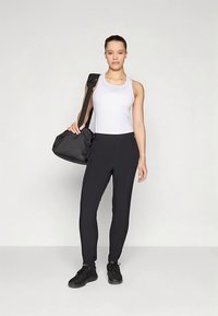 Ung kvinna klädd i vit linne, svarta byxor, svarta sneakers, bärande en svart gymväska över axeln, stående mot en enhetlig bakgrund.