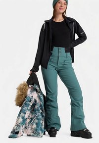 Türkiz snowboard nadrág magas derékkal és négy elülső gombbal, fekete cipzáras dzsekivel és színes mintás kabáttal, szőrmével díszítve.