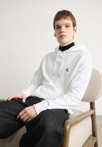 Sudadera blanca con un logotipo, cordones y bolsillo frontal; combinada con jeans de mezclilla oscura que muestran acentos de costura. Modelo sentado en una silla de madera.