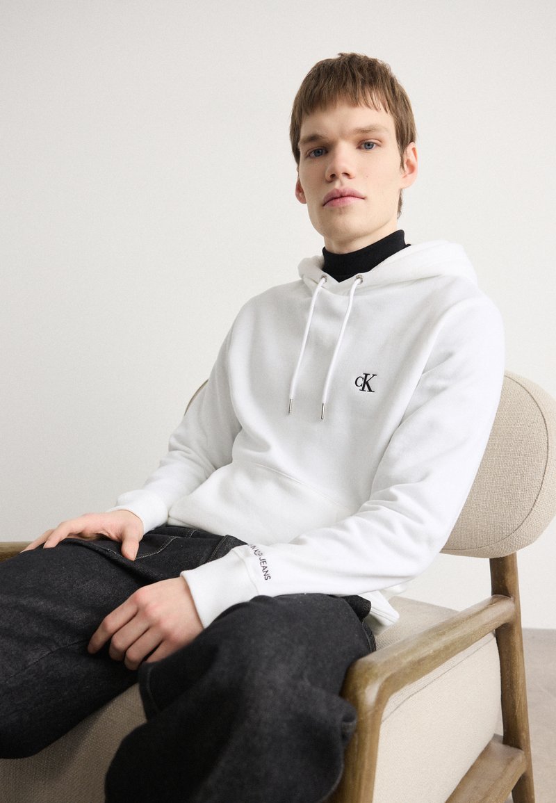 Sudadera blanca con un logotipo, cordones y bolsillo frontal; combinada con jeans de mezclilla oscura que muestran acentos de costura. Modelo sentado en una silla de madera.
