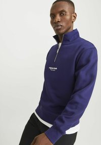 Sudadera de color azul marino con cremallera, fabricada en un tejido suave, que cuenta con un cuello alzado y un logo blanco en el pecho, combinada con una camiseta blanca debajo.