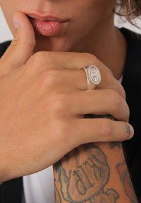 Silvrings med oval form som har en central ögon-design, texturerad band, bärs på en hand med synliga tatueringar. Neutral bakgrund.
