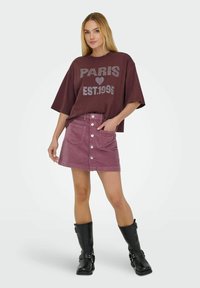 T-shirt bordeaux surdimensionné avec des lettres métalliques, associé à une mini-jupe en velours côtelé rose avec des poches avant et des boutons argentés, et des bottes noires à hauteur de genou.