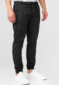 Black jogger pants with drawstring waist, elastic cuffs, and subtle branding on waistband. Paired with white sneakers for contrast.