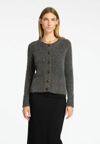 Cardigan in lana grigia con scollatura rotonda, maniche lunghe e sei bottoni marroni scuri sulla parte anteriore, abbinato a una gonna nera.