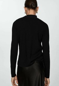 Femme avec les cheveux en chignon, portant de grandes boucles d'oreilles en or, un haut noir à manches longues et une jupe en satin noir, vue de dos.