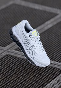 Sapatilha branca desportiva com parte superior texturizada, detalhes cinzentos e sola preta. Apresenta atacadores e um pequeno logótipo amarelo brilhante na língua.