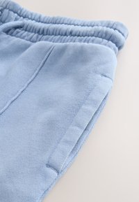 Pantalon de survêtement bleu clair en tissu texturé, avec une ceinture élastique et des poches latérales, cousu avec des coutures et des bords détaillés.
