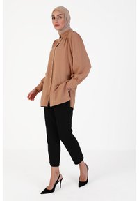 Blusa marrone chiaro con bottoni, maniche lunghe e tasche laterali, abbinata a pantaloni neri a sigaretta e tacchi slingback neri.