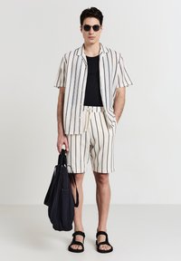 Homme portant des lunettes de soleil noires, une chemise noire, une chemise à manches courtes blanche à boutons et un short à rayures verticales noires, des sandales noires, tenant un sac noir.