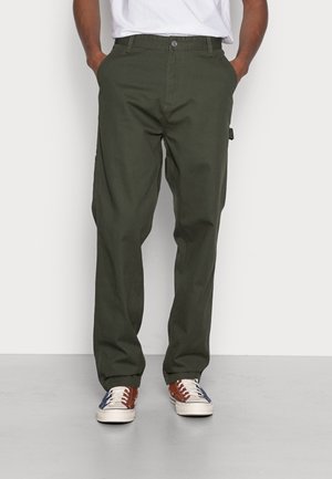 Mann trägt olivgrüne, gerade geschnittene Hose, Hände in den Taschen, und mehrfarbige Freizeitsneaker, steht auf grauem Boden.