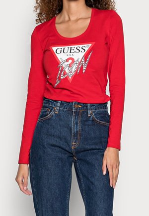 Kobieta ubrana w czerwony top z długim rękawem z logo "Guess Icon" oraz wysokim stanem niebieskich dżinsów, widoczna od szyi do bioder.