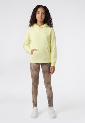 Gele hoodie met kangoeroezak, gecombineerd met bruine tie-dye legging. Model draagt witte sneakers. Soepele textuur, casual pasvorm.