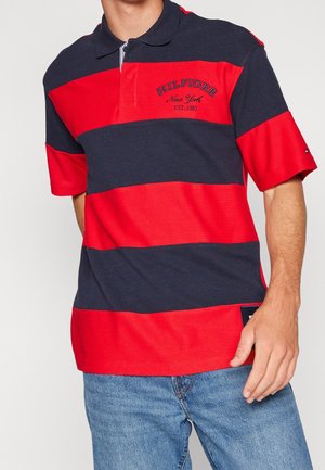 Mężczyzna w czerwono-granatowej koszulce polo w paski z haftem "Hilfiger New York Est. 1985", w parze z niebieskimi dżinsami.