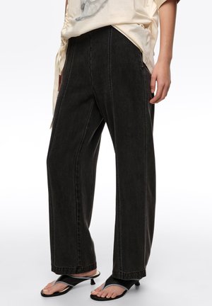 Mujer con jeans negros holgados con costuras verticales, una blusa beige clara y sandalias negras de tacón con tiras de entre los dedos.