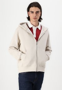 Light beige zip-up hoodie with a front pocket, adjustable hood, and ribbed hem. Worn over a red collared shirt and paired with blue jeans.