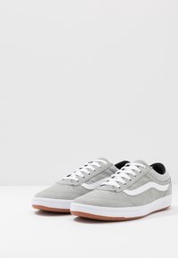 Baskets en daim gris avec des lacets blancs et une bande latérale, présentant une semelle intermédiaire blanche et une semelle extérieure en caoutchouc beige. Texture lisse et design discret.