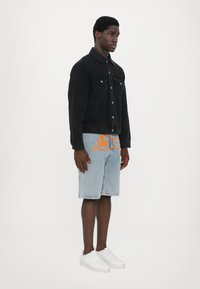 Veste en denim noire avec fermeture à boutons, deux poches poitrine, associée à un short bleu clair avec un design graphique orange et des baskets blanches.