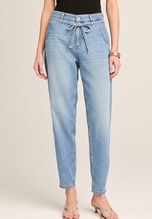 Frau trägt hellblaue Jeans mit hoher Taille und vorderer Bindegürtel, kombiniert mit einem schwarzen Oberteil und offenen Sandaletten mit Absatz.