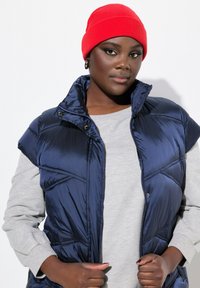 Gilet imbottito blu navy con colletto alto e bottoni a scatto, indossato sopra una felpa grigia. Il modello indossa un berretto lavorato a maglia rosso brillante.