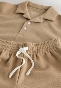 Polo en maille beige avec deux boutons blancs, associé à un pantalon assorti à taille élastique avec un cordon blanc.