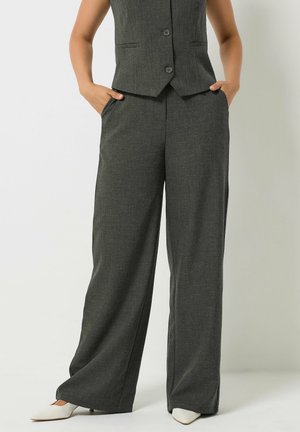 Femme portant un pantalon large gris foncé et un gilet boutonné assorti, les mains dans les poches, associé à des chaussures blanches pointues.