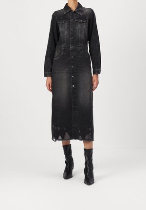 Kobieta w wyblakłej czarnej midi sukience z denimu z guzikami, z długimi rękawami i postrzępionym dołem, w połączeniu z czarnymi skórzanymi botkami na obcasie sięgającymi do połowy łydki.