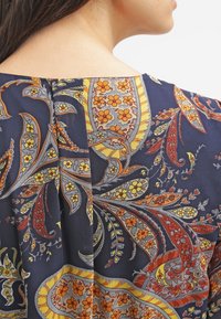 Det blå klädestyget har ett färgglatt paisleymönster med gula och oranga blommor. Urrigningen har en subtil veckdetalj.