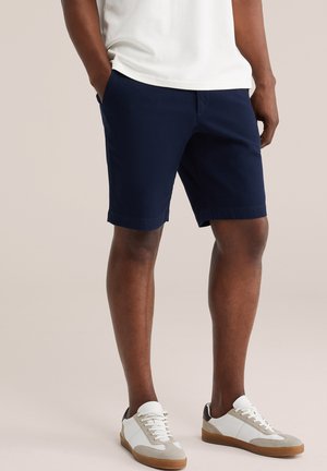 Man draagt marineblauwe korte broek, witte casual sneakers en een wit shirt met handen in de zakken tegen een effen achtergrond.