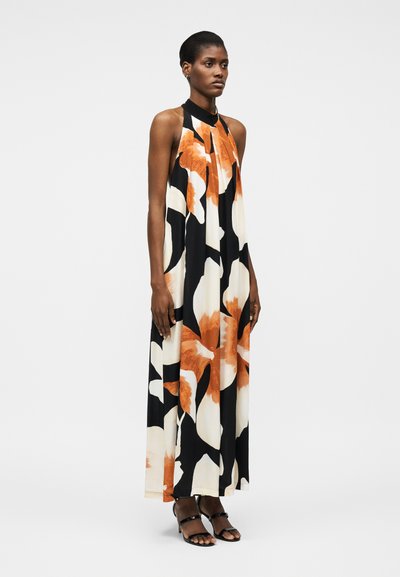 Γυναίκα όρθια που φορά αμάνικο, μακρύ φόρεμα με halter λαιμόκοψη και μεγάλο πορτοκαλί και λευκό λουλουδάτο μοτίβο, σε συνδυασμό με μαύρα πέδιλα με τακούνι.