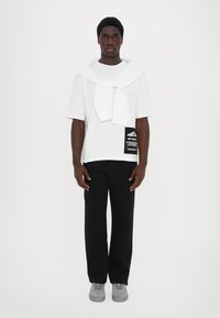 Weißes T-Shirt mit kurzen Ärmeln und schwarzem Label, kombiniert mit schwarzen Hosen und grauen Sneakern. Ein weißer Pullover liegt über den Schultern.