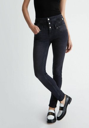 Frau trägt schwarze Skinny-Jeans mit hoher Taille und Knopfdetail sowie zweifarbige schwarz-weiße Loafers, steht mit einer Hand in der Tasche.