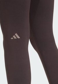 Leggings sportivi neri con una texture liscia, caratterizzati da un discreto logo Adidas marrone sulla coscia e dettagli di cuciture rinforzate.