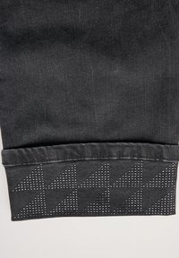 Pantalón de mezclilla negro con un dobladillo que presenta un patrón triangular de tachuelas en plata. La textura es lisa con un ligero brillo.