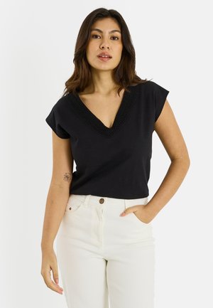 Mujer vistiendo una blusa negra de manga corta con escote en V y detalles de encaje, pantalones blancos, parada con una mano en el bolsillo frente a un fondo blanco.