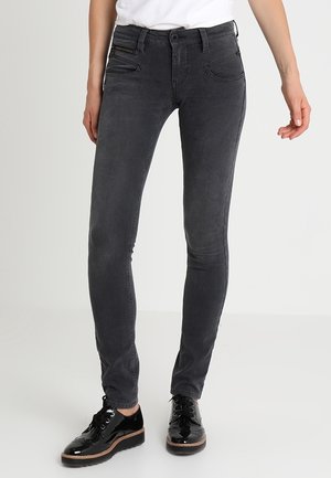 Schmal geschnittene schwarze Jeans, getragen von einer Person, kombiniert mit glänzenden schwarzen Schnürschuhen und einem weißen, in die Hose gesteckten Oberteil, stehend vor einem einfarbigen Hintergrund.