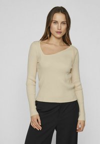 Top beige a maniche lunghe a costine con scollatura asimmetrica e vestibilità slim, indossato con pantaloni neri. La texture sembra morbida ed elastica.