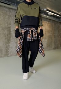 Olivgrön och svart sweatshirt med tre ränder, kombinerad med lösa svarta byxor och en brun rutig skjorta bunden i midjan. Vita sneakers.