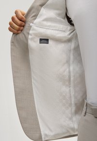 Une main tient ouvert une veste de costume beige, révélant la doublure intérieure blanche à motifs avec une petite étiquette indiquant "SUPER 100'S NATURAL STRETCH".