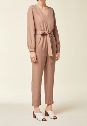 Femme portant une combinaison-pantalon cache-cœur à manches longues rose poudré avec ceinture nouée et sandales blanches à talons blocs et bouts ouverts.