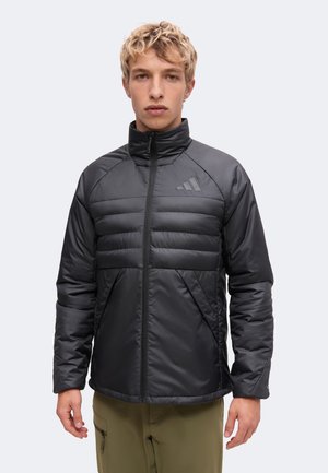 Junger Mann mit blondem lockigem Haar, der eine schwarze wattierte Jacke mit Reißverschluss und olivgrüne Hose trägt, steht vor einem einfarbigen Hintergrund.