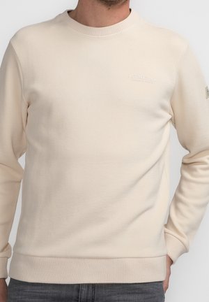 Mężczyzna ubrany w kremowy sweter z dekoltem typu crew neck, wykonany z subtelnej teksturowanej tkaniny, z małym logo na piersi, w połączeniu z ciemnoszarymi dżinsami.