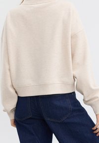 Beige kort sweatshirt med texturerad tyg, vida ärmar och rundad halsringning, tillsammans med mörka denimbyxor som har bakfickor.