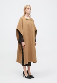 Καφέ μάλλινο πόντσο με χαλαρή εφαρμογή, διαθέτοντας στρογγυλή λαιμόκοψη, πλευρικές σχισμές και διακοσμητικό κούμπωμα μπροστά. Φοριέται πάνω από ένα σκούρο φόρεμα.