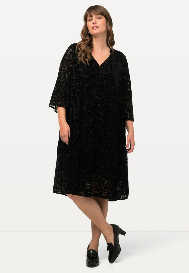 Abito nero di velluto con sovrapposizione in pizzo floreale, scollo a V, maniche a tre quarti e vestibilità ampia, con tasche laterali e pinces.