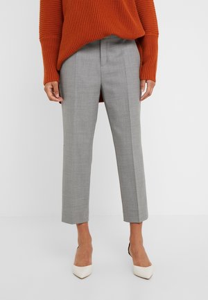 Vrouw draagt grijze, op maat gemaakte enkelhoge broek, roestoranje gebreide trui en witte pumps met puntige neus tegen een effen witte achtergrond.