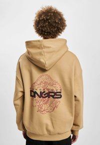 Sudadera oversized color caqui con un gráfico de dragón rojo y texto negro "DANGEROUS DNGRS" en la parte trasera. Tela suave, mangas largas y capucha.