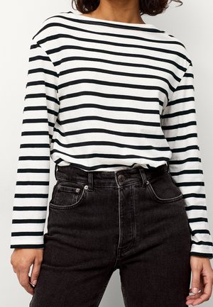 Woman wearing a long-sleeve white shirt with black horizontal stripes tucked into high-waisted black jeans with pockets.